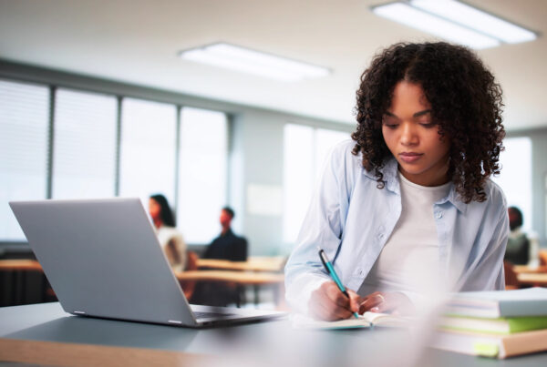 Student sitting at a desk in front of a laptop writing on a piece of paper with a pencil.