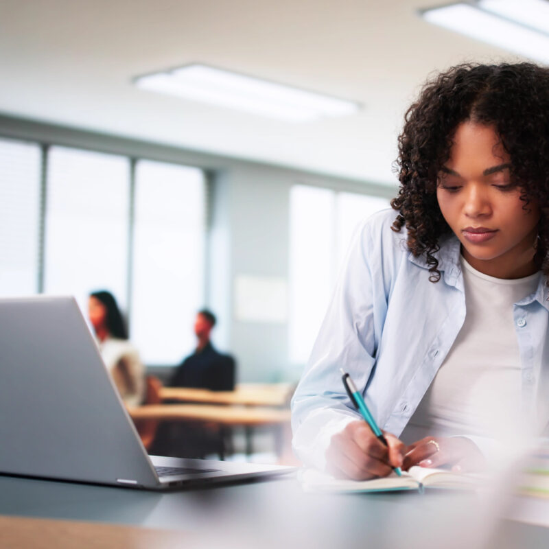 Student sitting at a desk in front of a laptop writing on a piece of paper with a pencil.