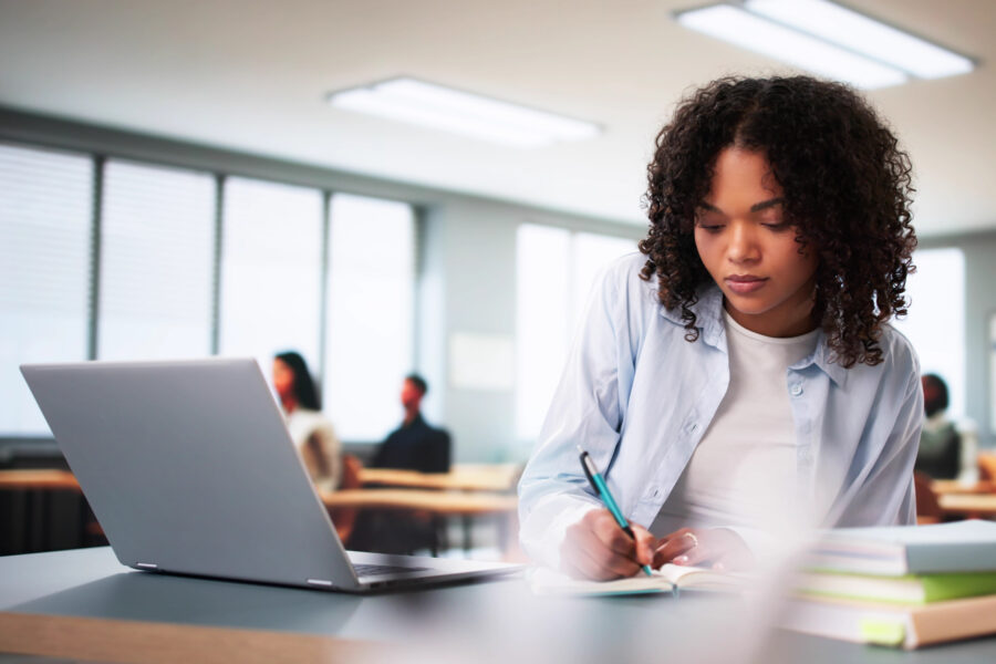 Student sitting at a desk in front of a laptop writing on a piece of paper with a pencil.